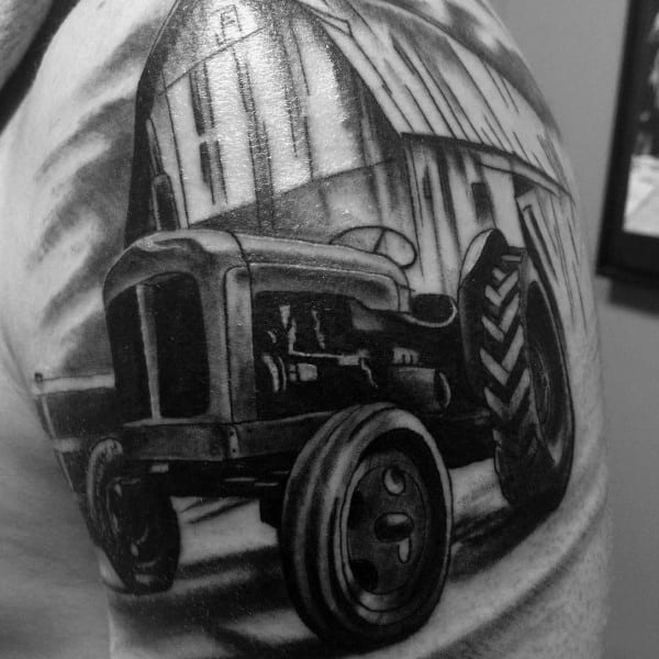 Bold black and gray upper arm tattoo of a vintage tractor in front of a large barn, with strong shading and perspective.