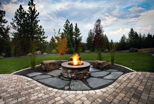 A cozy stone fire pit on a patio, enveloped by trees and grass beneath a partly cloudy sky