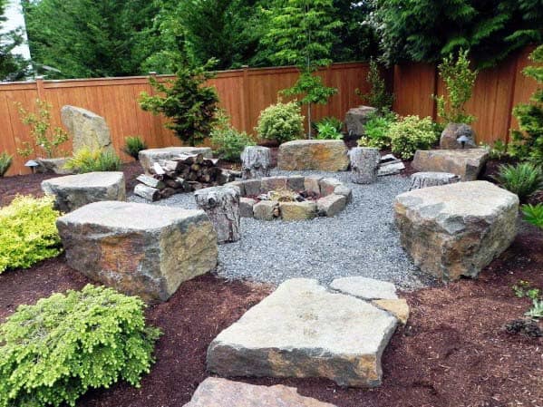 Stone seating surrounds a fire pit in a landscaped yard with lush greenery and a wooden fence