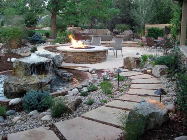 Stone path leading to a fire pit with chairs, surrounded by landscaping and a small waterfall feature
