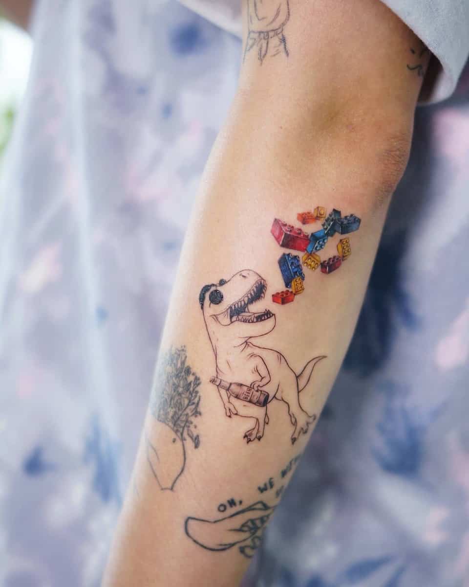 Playful forearm tattoo featuring a tiny T-rex holding a bottle, with colorful LEGO bricks floating above, adding whimsy and fun