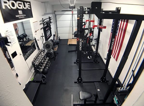 Compact garage gym with Rogue squat rack, dumbbell rack, weight plates, and rubber flooring.