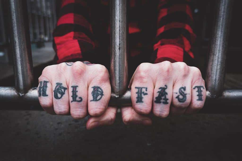 Hands gripping bars with "HOLD" and "FAST" tattooed on the knuckles