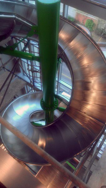 A large, indoor spiral slide of shiny metal hues gleams with vibrant green supports