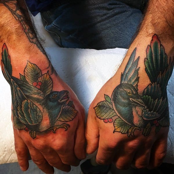 Traditional-style magpie tattoos on both hands, featuring bold black, blue, and green feathers with red berry accents for a striking, symmetrical design.