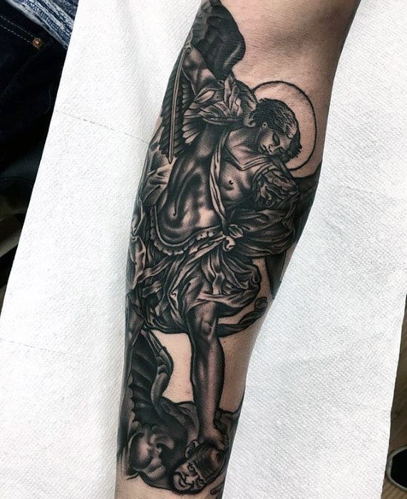A detailed black and grey tattoo on the forearm featuring an angel with large wings, holding a sword, and defeating a fallen figure beneath.