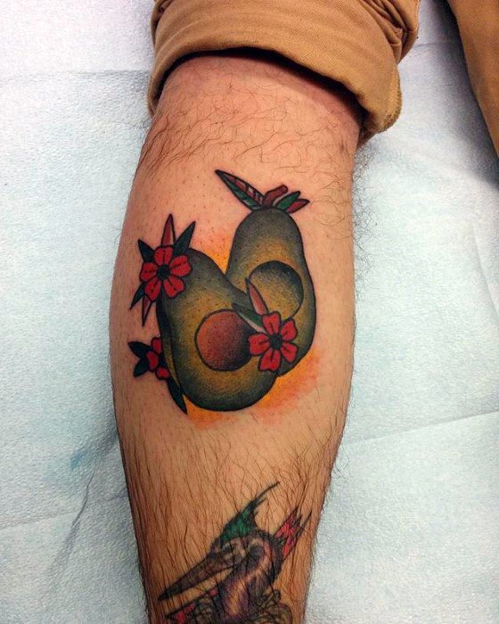 Traditional-style tattoo of two avocado halves forming a heart shape, decorated with red flowers and soft orange shading on the calf.
