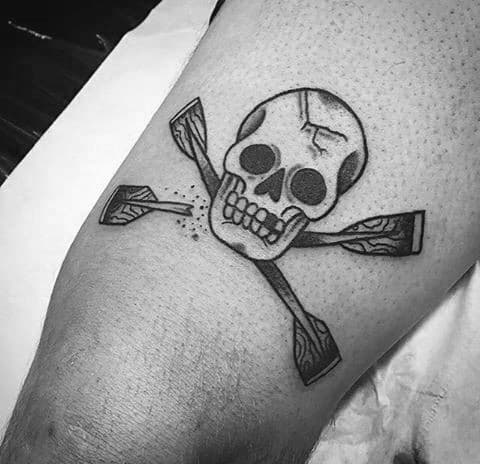 Alt text: Bold black and grey tattoo of a skull with crossed canoe paddles beneath it, resembling a pirate-inspired design.