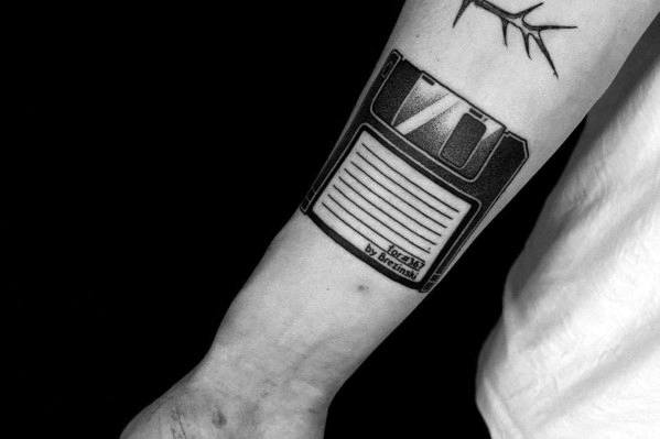 Bold black and grey tattoo of a detailed floppy disk on the inner forearm, featuring realistic shading and vintage label text.