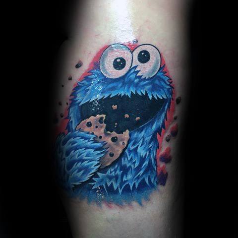 Vibrant Cookie Monster tattoo with excited expression, holding a bitten cookie as crumbs fly, set against a red and black splash background.