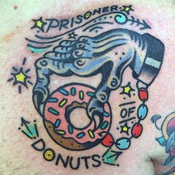 Tattoo of a skeleton hand holding a pink donut with the phrase prisoner of donuts, surrounded by stars, beads, and playful arrows.