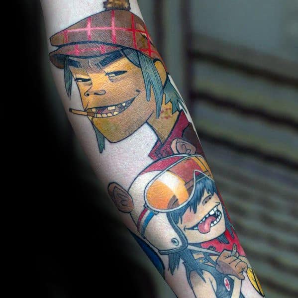 Vibrant forearm tattoo of Murdoc grinning with a cigarette and Noodle wearing a bear helmet, sticking her tongue out in a playful pose.