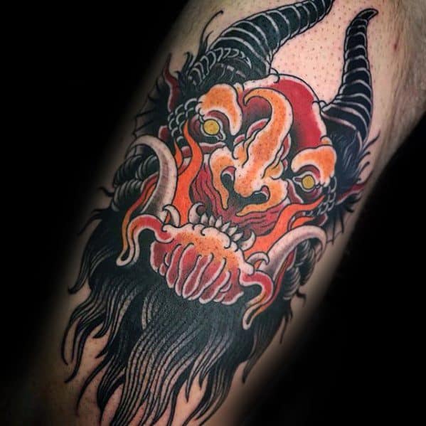 Fiery Krampus tattoo with a fierce red and orange demon face, large tusks, glowing yellow eyes, black horns, and flowing dark beard.