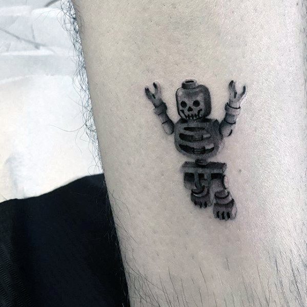 Small black and grey tattoo of a Lego skeleton minifigure with raised arms, detailed with strong shadows for a 3D and playful look.