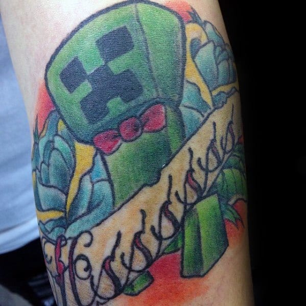 Colorful Minecraft Creeper tattoo wearing a red bow tie, surrounded by flowers and a scroll banner with decorative script.