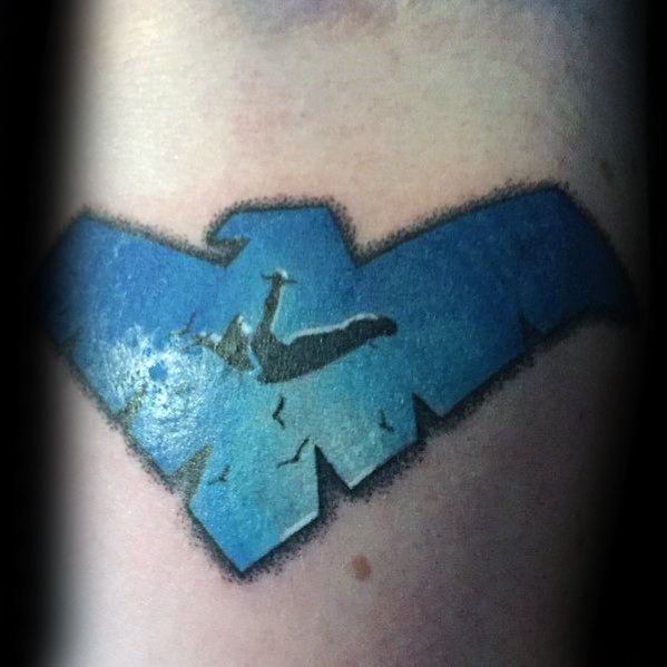 Nightwing emblem tattoo with a silhouette of him free-falling among birds, set inside a blue stylized wing symbol.