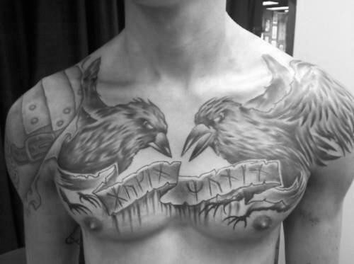 Black and grey chest tattoo of Huginn and Muninn facing each other over a torn scroll with runes, spreading across both pectorals.