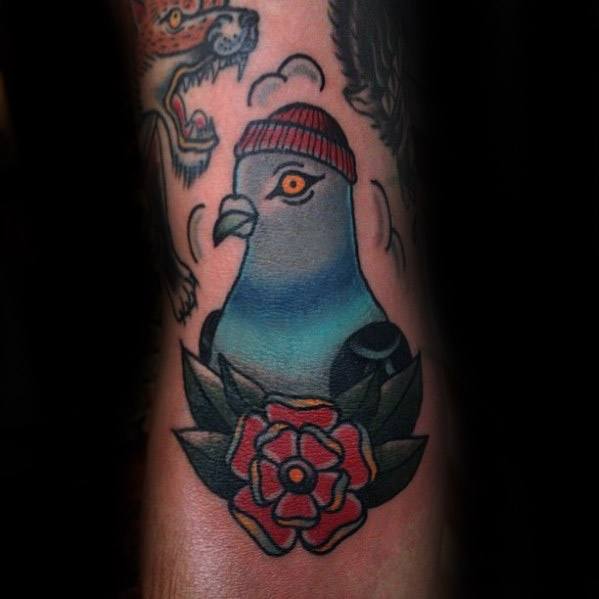 Bold traditional pigeon tattoo with a red beanie and classic rose, combining street personality and timeless tattoo symbolism.