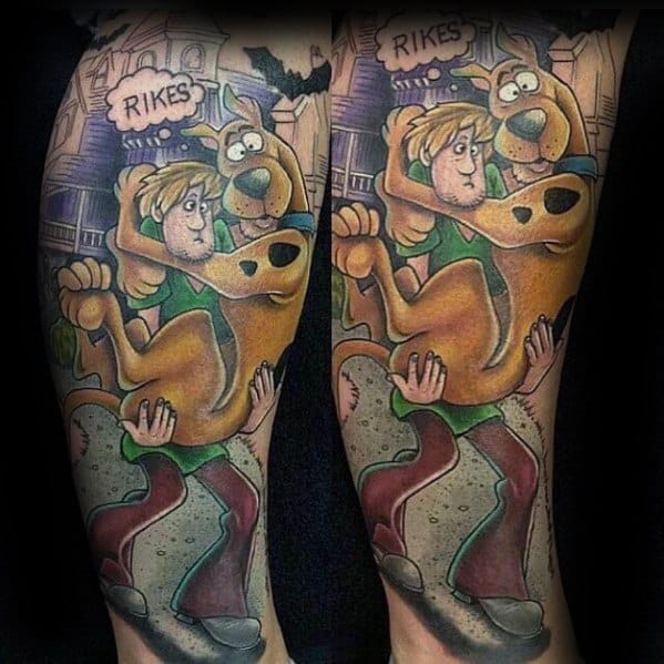 Tattoo of Scooby-Doo jumping into Shaggy’s arms in fear, with a haunted house background and “RIKES” in a speech bubble above.