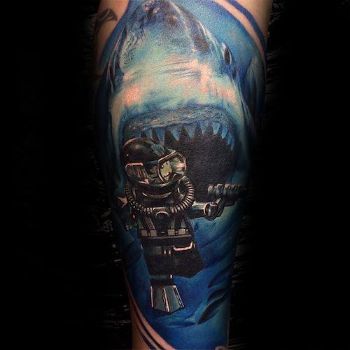 Colorful tattoo of a LEGO scuba diver in front of a massive, realistic shark with an open mouth in deep blue ocean waters.