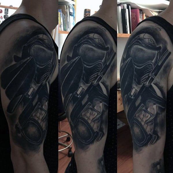 Realistic black and grey tattoo of a paintball player in full gear, featuring a helmet, goggles, and marker gun on the upper arm.