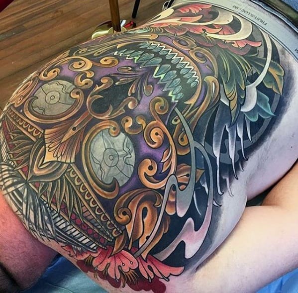 Colorful Tibetan skull tattoo on the upper thigh and hip with fierce teeth, vibrant swirling patterns, and ornate golden detailing.