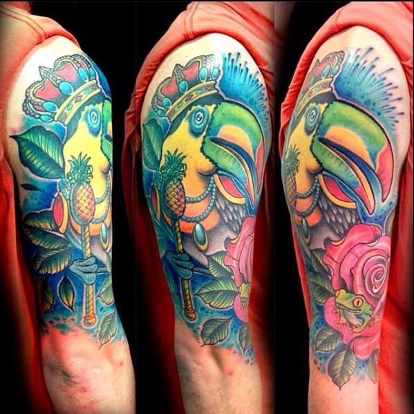 Vibrant full upper arm toucan tattoo with a royal theme, featuring a crown, scepter with a pineapple, and colorful tropical flowers.