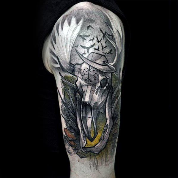 Artistic Witcher tattoo on the upper arm featuring a stylized Leshen skull with antlers, yellow highlights, runes, and flying birds.
