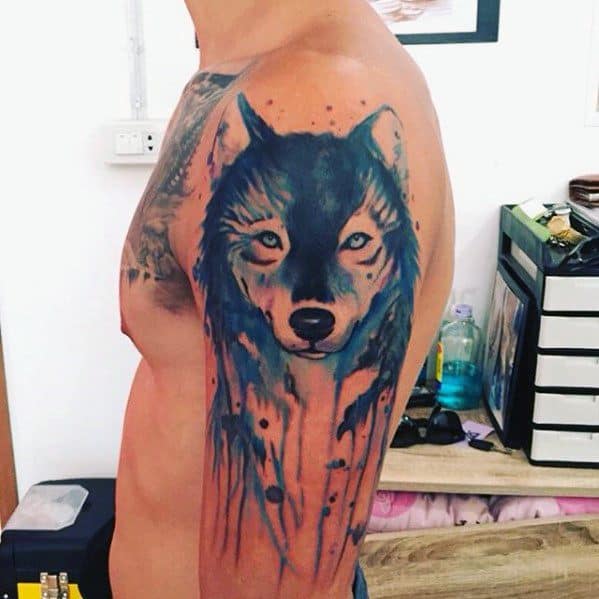 Blue watercolor wolf tattoo on upper arm with a calm, focused gaze and dripping paint effect for a bold and artistic statement.