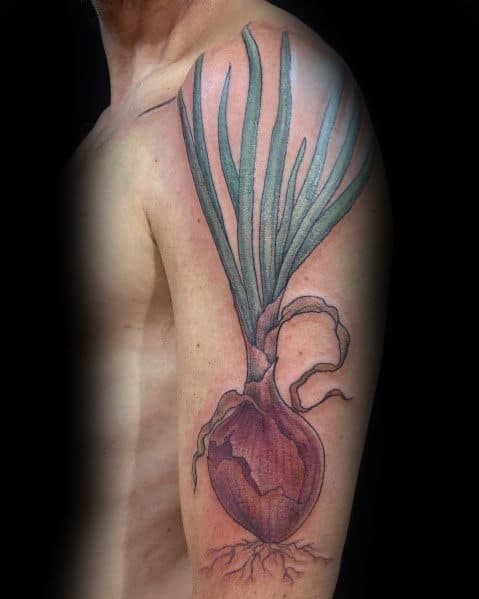 Realistic onion tattoo on the upper arm, featuring detailed shading, long green leaves, and exposed roots for a natural botanical design.