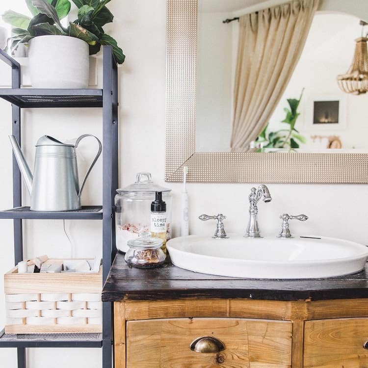Farmhouse bathroom sink with a wooden vanity, plants, and a mirror; a watering can sits on a shelf