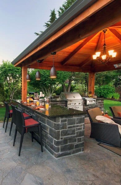 Outdoor stone bar with a wooden pavilion, pendant lighting, and a built-in grill on a stone patio.