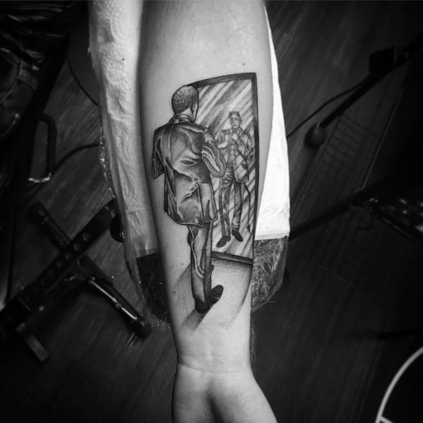 Tattoo of a man in a suit looking into a mirror, with his reflection appearing slightly altered, creating a surreal and thought-provoking design.