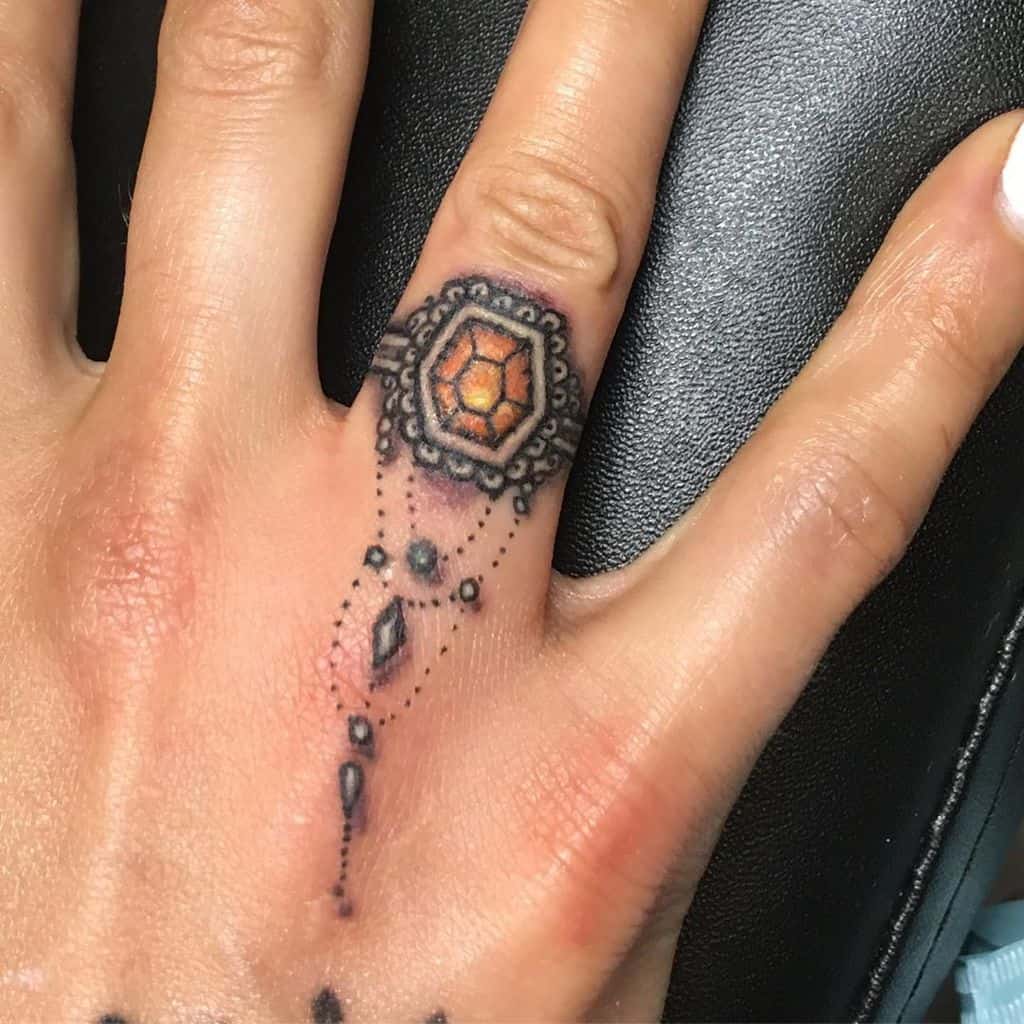 Ornate gemstone ring tattoo, featuring an orange gem with intricate black and gray dot patterns and embellishments