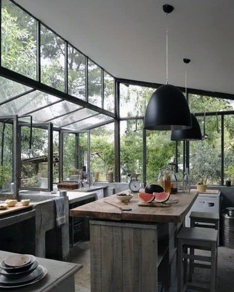 Rustic kitchen with wooden island, glass walls, black pendant lights, and lush outdoor views.
