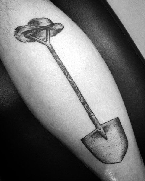 Black and grey tattoo of a detailed shovel with a long handle, wrapped in a ribbon, symbolizing hard work, perseverance, and resilience.