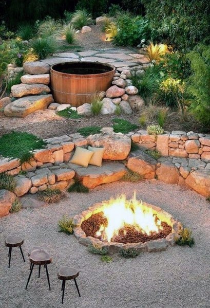 Outdoor seating area with a fire pit, stools, and a hot tub surrounded by stones and greenery