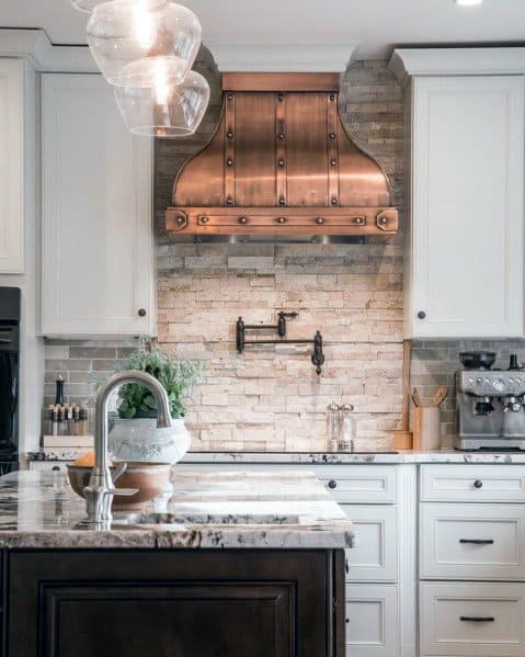 Rustic and luxurious kitchen featuring a copper range hood, textured stone backsplash, and elegant white cabinetry. A marble island countertop adds sophistication to the space, complemented by modern fixtures and decor accents.