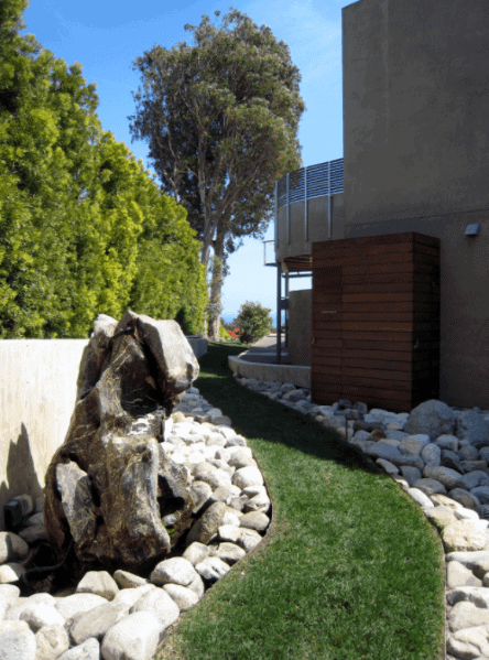A modern garden pathway bordered with large stones, featuring a grassy path and a large rock feature, with lush greenery surrounding the space