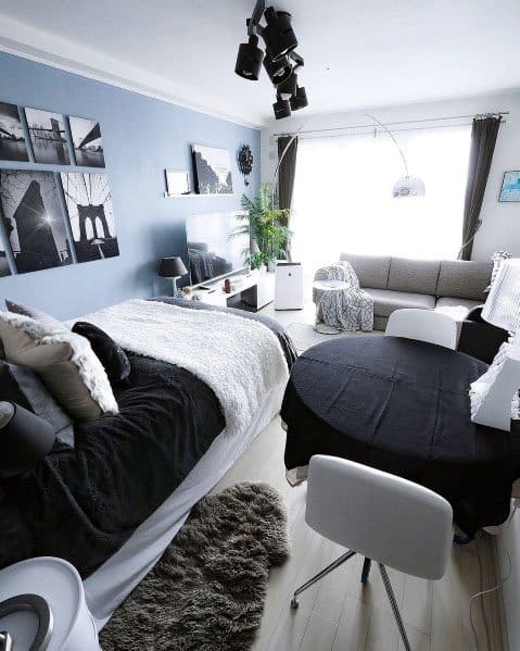 Modern studio apartment with a blue accent wall, cozy bedding, black dining setup, and bright natural light.