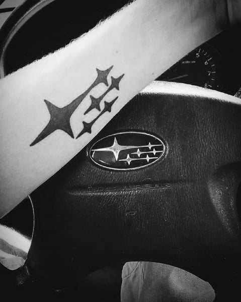 Black Subaru logo tattoo on the forearm, featuring the iconic six-star emblem, shown alongside the matching Subaru steering wheel badge.
