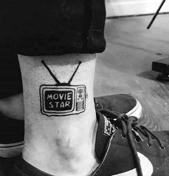 Minimalist black ink tattoo of a vintage TV on the ankle, displaying the words "MOVIE STAR" on the screen, with simple bold lines and classic antennas.