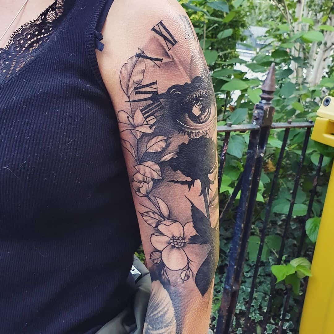Upper arm tattoo with eye, clock, and floral design in black ink