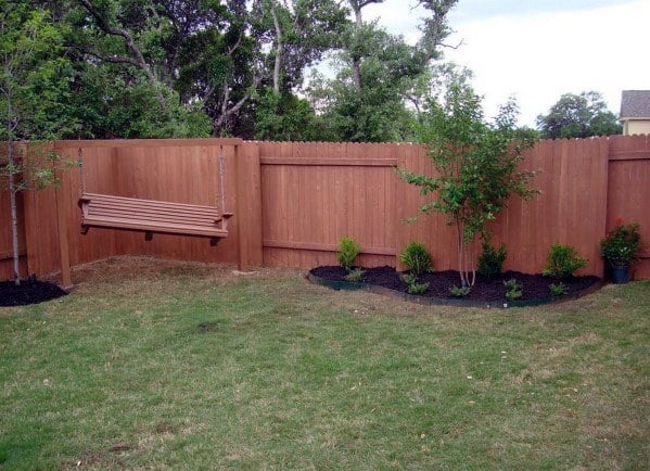 Wooden fence with a hanging bench, amidst trees and shrubs, grace the green yard
