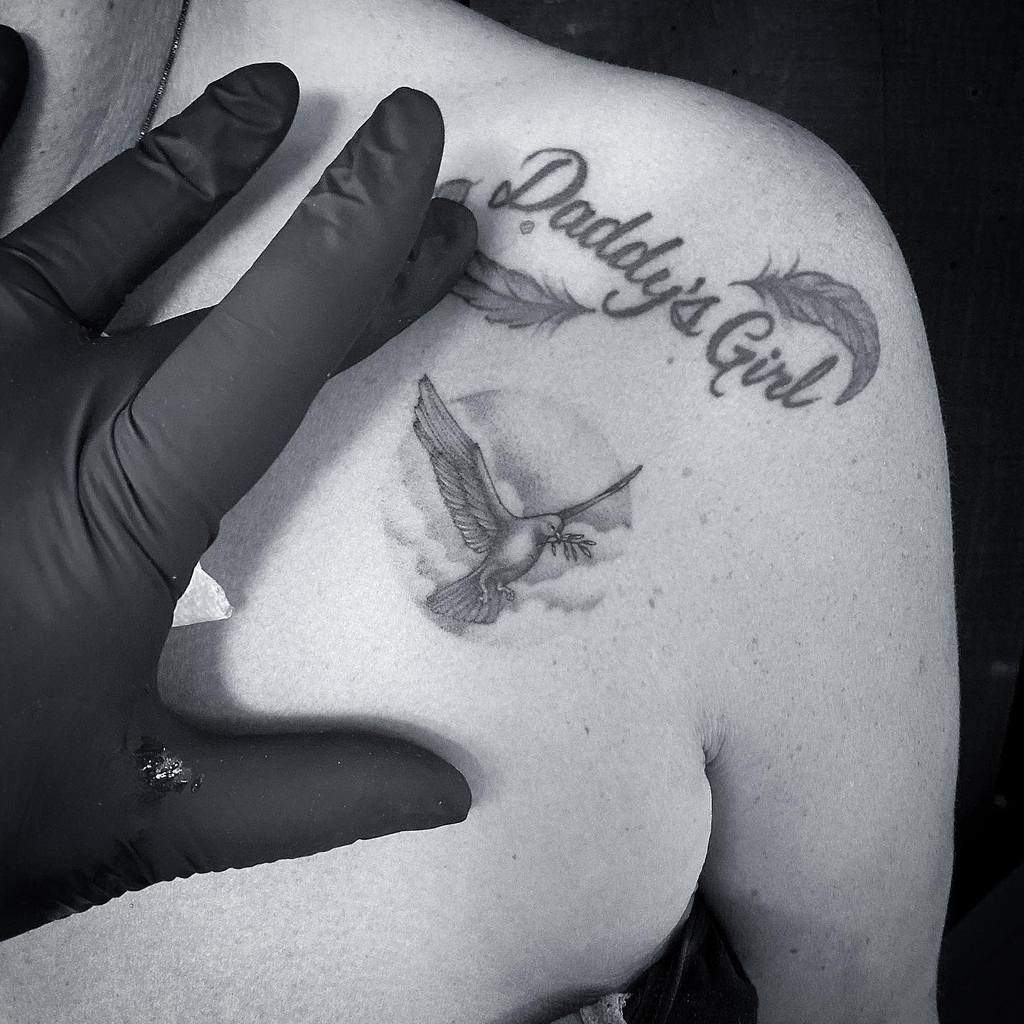 Gloved hand near a stunning single needle shoulder tattoo of a bird and the text "Daddy's Girl" with two delicate feathers