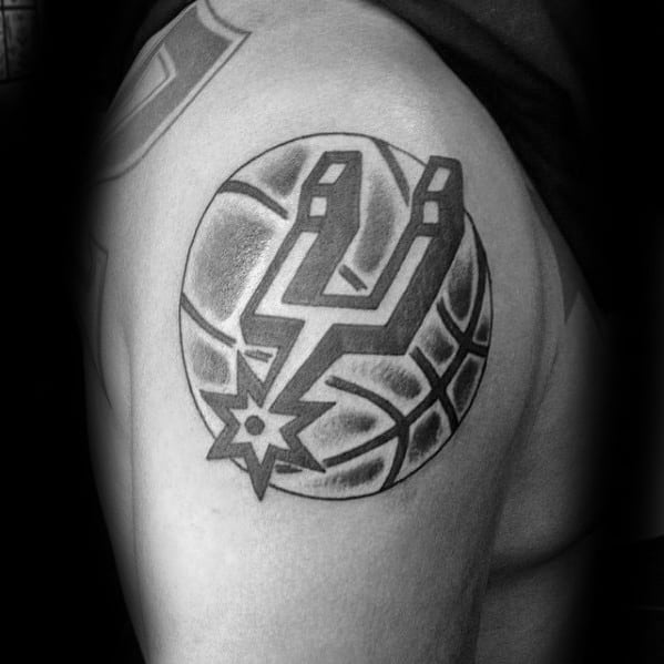 Black and gray San Antonio Spurs logo tattoo placed over a shaded basketball design, inked on the upper arm with bold lines and contrast.