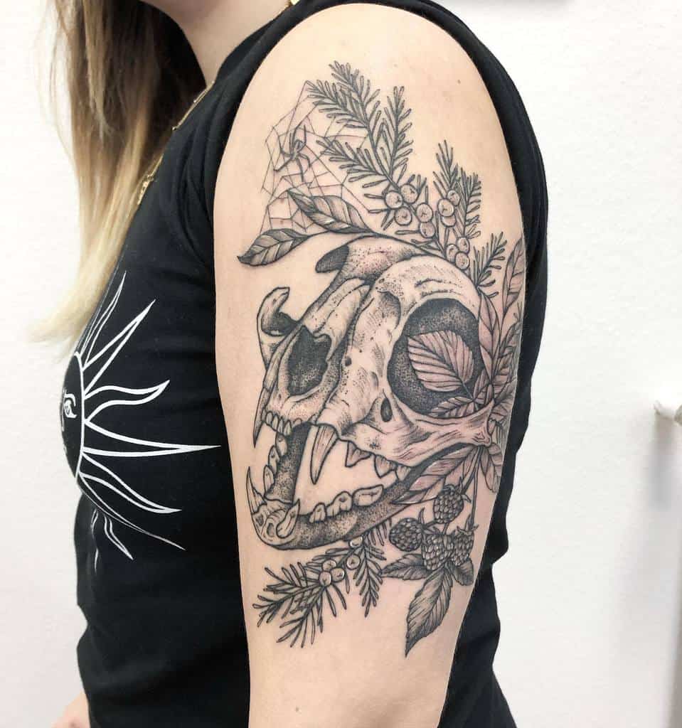 An upper arm tattoo featuring a detailed cat skull surrounded by botanical elements, including leaves and berries