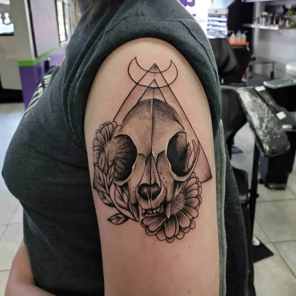 An upper arm tattoo featuring a geometric cat skull design with crescent moon symbols and flowers on both sides