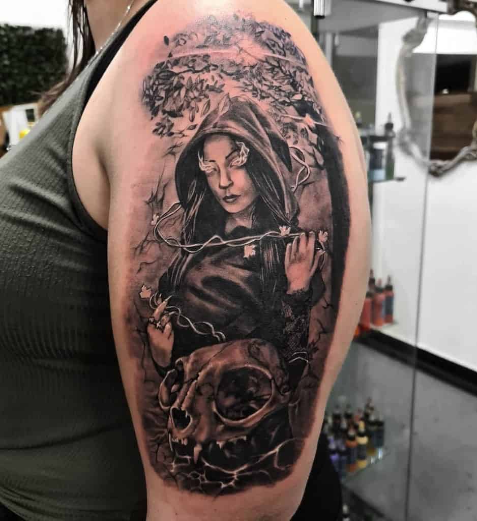 A detailed upper arm tattoo featuring a hooded woman with flowers covering her eyes, vines wrapped around her, and a large cat skull at the bottom, set against a mystical background