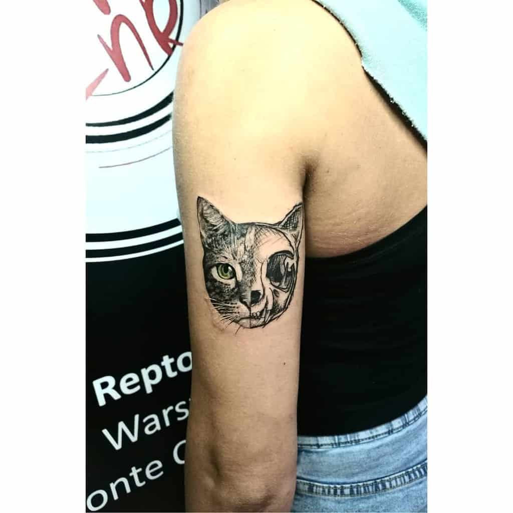 An upper arm tattoo featuring a split image of a cat's face, with one side showing a realistic cat and the other half a cat skull